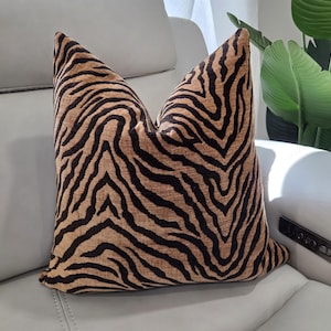 May include: A square throw pillow featuring a textured fabric with a black and tan zebra stripe pattern. The pillow has a plump, filled appearance with defined corners and a consistent, repeating animal print design across the front surface.