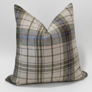 May include: A square throw pillow featuring a plaid pattern in shades of cream, olive green, and muted blue. The fabric has a visible woven texture with intersecting horizontal and vertical lines forming a classic tartan-inspired grid design.