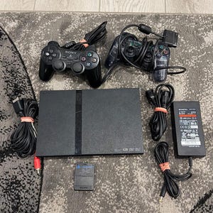May include: A black Sony PlayStation 2 game console with two controllers, one black and one translucent blue, a black memory card, a power adapter, and various connection cables. The console features a rectangular design with a central disc tray.