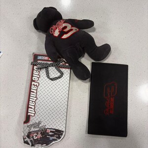 Può includere: Un orsacchiotto di peluche nero che indossa una maglietta con il numero 3, un espositore rettangolare NASCAR di Dale Earnhardt con motivo a piastra diamantata e nastro a scacchi, e una custodia rettangolare nera con il numero 3 rosso e il logo NASCAR.
