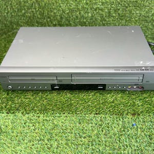 May include: A silver DVD and VHS player combo unit featuring a front-loading disc tray on the left and a cassette slot on the right. The device includes a power button, multiple control buttons, a central display panel, and front-facing input ports.