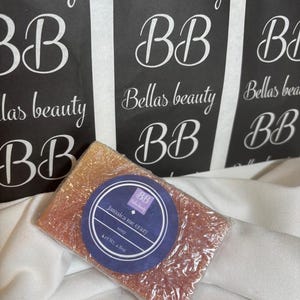 Könnte beinhalten: Ein rechteckiges Stück Seife mit schimmernder goldener und rosafarbener Textur, verpackt in Klarsichtfolie. Ein rundes violettes Etikett trägt den Text "BB Bellas beauty", "Jamaica me crazy", "soap" und "Net Wt. 136g". Schwarze Etiketten sind im Hintergrund.