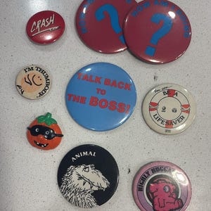 Op de afbeelding: Een verzameling van negen ronde buttons met diverse ontwerpen, waaronder teksten zoals CRASH, HOW AM I DOING, TALK BACK TO THE BOSS, I'M THUMBODY, BE A LIFESAVER, ANIMAL en HIGHLY HUGGABLE, naast een kleine oranje pompoenvormige speld.