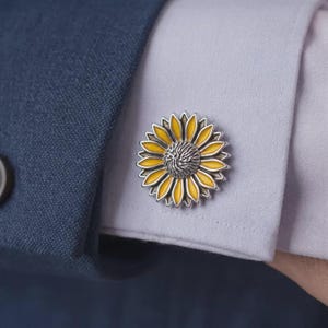 Może przedstawiać: Srebrzysta spinka do mankietu w kształcie słonecznika z płatkami z jasnej żółtej emalii i teksturowanym, ciemnosrebrnym środkiem. Słonecznikowa spinka do mankietu jest przymocowana do mankietu jasnoliliowej koszuli pod granatową marynarką.