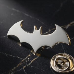 May include: A metal lapel pin featuring a white bat-shaped emblem with gold-toned edges. The pin includes a circular metal clutch backing with a post mechanism, designed for attachment to fabric or other materials, showcasing a clean and polished finish.