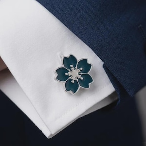 以下が含まれることがあります： 5枚の花びらを持つ花をかたどった銀色のカフスボタンで、各花びらには深いティールブルーのエナメルが施されています。カフスボタンは、濃紺のブレザーの袖の下に見える、パリッとした白いシャツの袖口のボタンホールに通されています。