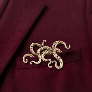 Puede incluir: Un broche de metal dorado con forma de pulpo con múltiples tentáculos curvados, que presenta una superficie texturizada con detalles de ventosas visibles, prendido en el bolsillo de una americana de color burdeos profundo con tela lisa.