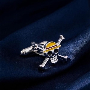 May include: A silver-toned metal cufflink featuring a skull and crossbones design. The skull wears a yellow hat with a red band. The eye sockets and nose are filled with black enamel, and the teeth are detailed with silver-toned metal lines.