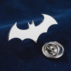 May include: A silver-toned metal lapel pin shaped like a stylized bat silhouette. The pin features a smooth, reflective surface and includes a circular metal clutch backing with a small central hole, designed for secure attachment to fabric garments.