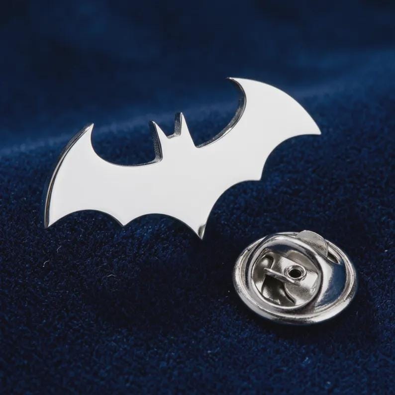 May include: A silver-toned metal lapel pin shaped like a stylized bat silhouette. The pin features a smooth, reflective surface and includes a circular metal clutch backing with a small central hole, designed for secure attachment to fabric garments.