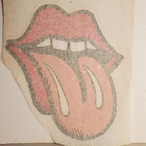 May include: A vintage, stylized image of a mouth with a protruding tongue. The lips are a vibrant red with a black outline, and the tongue is a lighter shade of red. The design is on a piece of aged paper, suggesting a retro aesthetic.