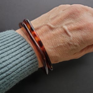 May include: Two brown and amber-colored bangles are stacked on a wrist. The bangles have a tortoiseshell pattern and a smooth, glossy finish. The wrist is wearing a teal-colored sweater.