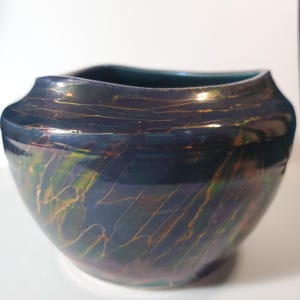 May include: A dark blue ceramic bowl with a unique iridescent finish. The bowl has a wide, slightly curved rim and a rounded body. Gold-colored lines streak across the surface, adding a decorative element to the piece.