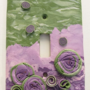 May include: A light switch cover made of purple and green clay with a marbled effect. The cover is decorated with swirls and flowers.