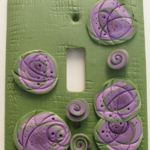 May include: Green textured light switch cover with purple and green spiral designs. The cover has a rectangular shape with a switch opening and is decorated with several raised, circular, and spiral-shaped embellishments.
