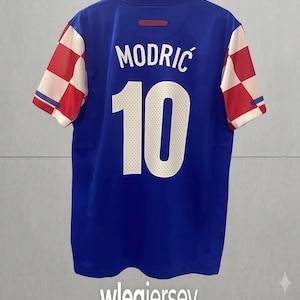 May include: A blue sports jersey featuring the name MODRIĆ and the number 10 in white on the back. The sleeves display a red and white checkered pattern. The jersey is displayed on a hanger against a plain background with the text wlegjersey below.