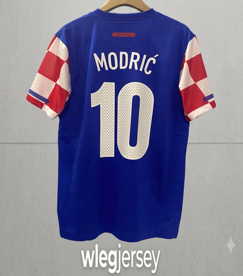 May include: A blue sports jersey featuring the name MODRIĆ and the number 10 in white on the back. The sleeves display a red and white checkered pattern. The jersey is displayed on a hanger against a plain background with the text wlegjersey below.
