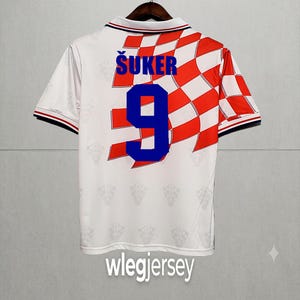 May include: A white short-sleeved jersey featuring a red and white checkered pattern on the right side. The back displays the name &Scaron;UKER in blue letters above the number 9 in blue. The fabric has a subtle repeating emblem pattern throughout.