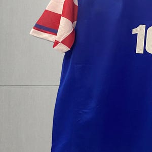 May include: A blue short-sleeved jersey featuring a white number 10 on the front and a red and white checkered pattern on the sleeve. The garment has a round neckline and is displayed on a wooden hanger against a plain light gray background.