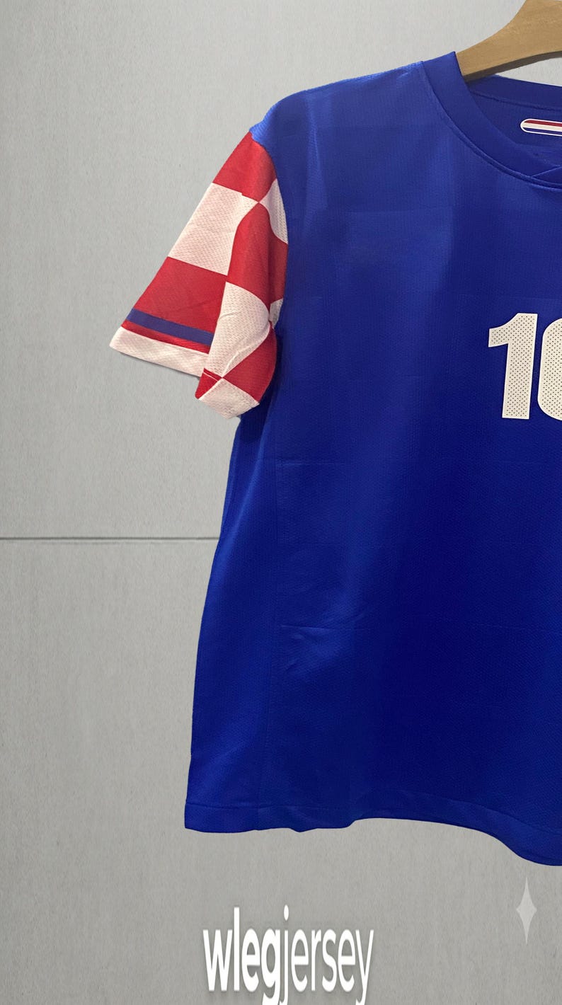 May include: A blue short-sleeved jersey featuring a white number 10 on the front and a red and white checkered pattern on the sleeve. The garment has a round neckline and is displayed on a wooden hanger against a plain light gray background.
