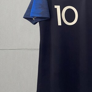 May include: A dark navy blue athletic jersey featuring the number 10 in white on the front. The short sleeves have a textured blue pattern, and the neckline includes a small red, white, and blue detail. The text wlegjersey is visible.