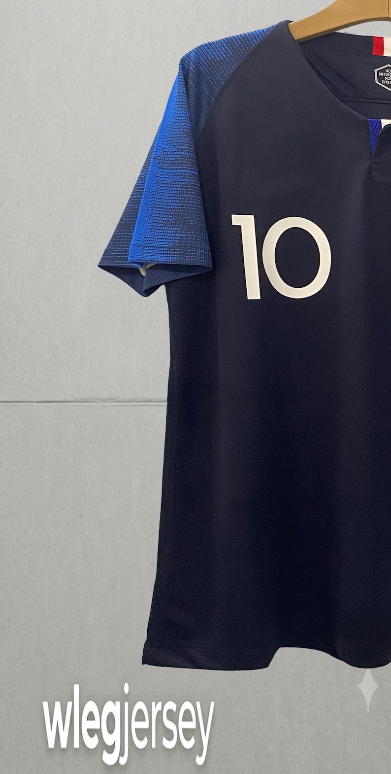 May include: A dark navy blue athletic jersey featuring the number 10 in white on the front. The short sleeves have a textured blue pattern, and the neckline includes a small red, white, and blue detail. The text wlegjersey is visible.