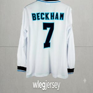 Puede incluir: Una camiseta deportiva blanca de manga larga con el nombre BECKHAM y el número 7 en negro con bordes azul claro en la parte posterior. El cuello y los puños son negros con ribetes azul claro. El texto wlegjersey aparece en la parte inferior.