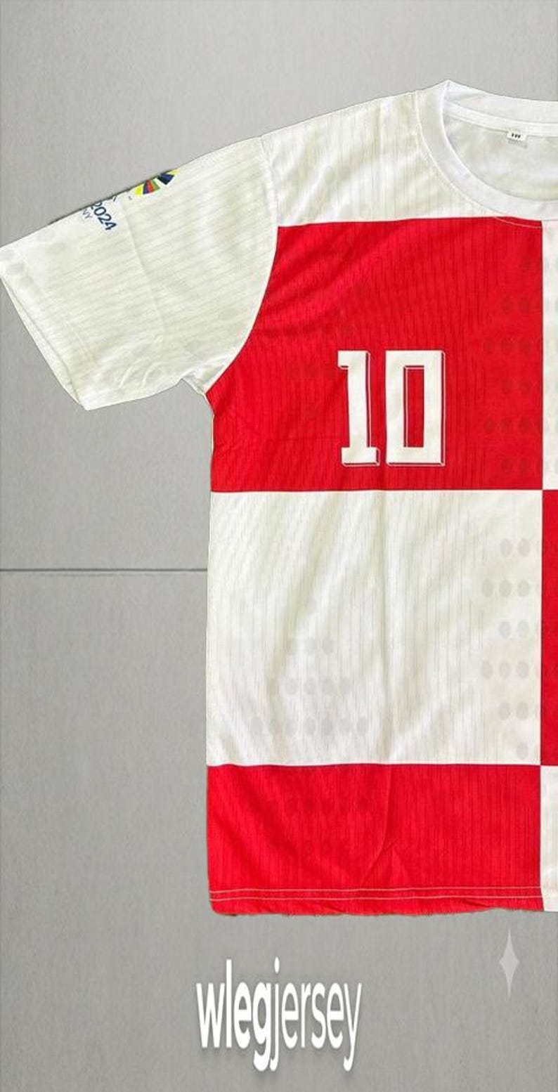 Pu&ograve; includere: Una maglia a maniche corte con un motivo a scacchi rossi e bianchi sul davanti con il numero 10 stampato in bianco. La manica include una piccola grafica colorata e il testo 2024. La parola wlegjersey &egrave; stampata nella parte inferiore.