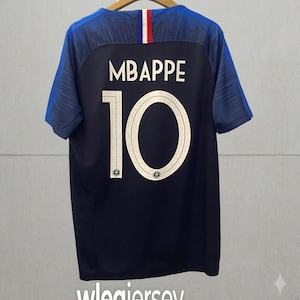 May include: A dark blue short-sleeved sports jersey featuring the name MBAPPE and the number 10 in white on the back. The collar area includes a vertical blue, white, and red stripe detail. The sleeves have a textured pattern in shades of blue.