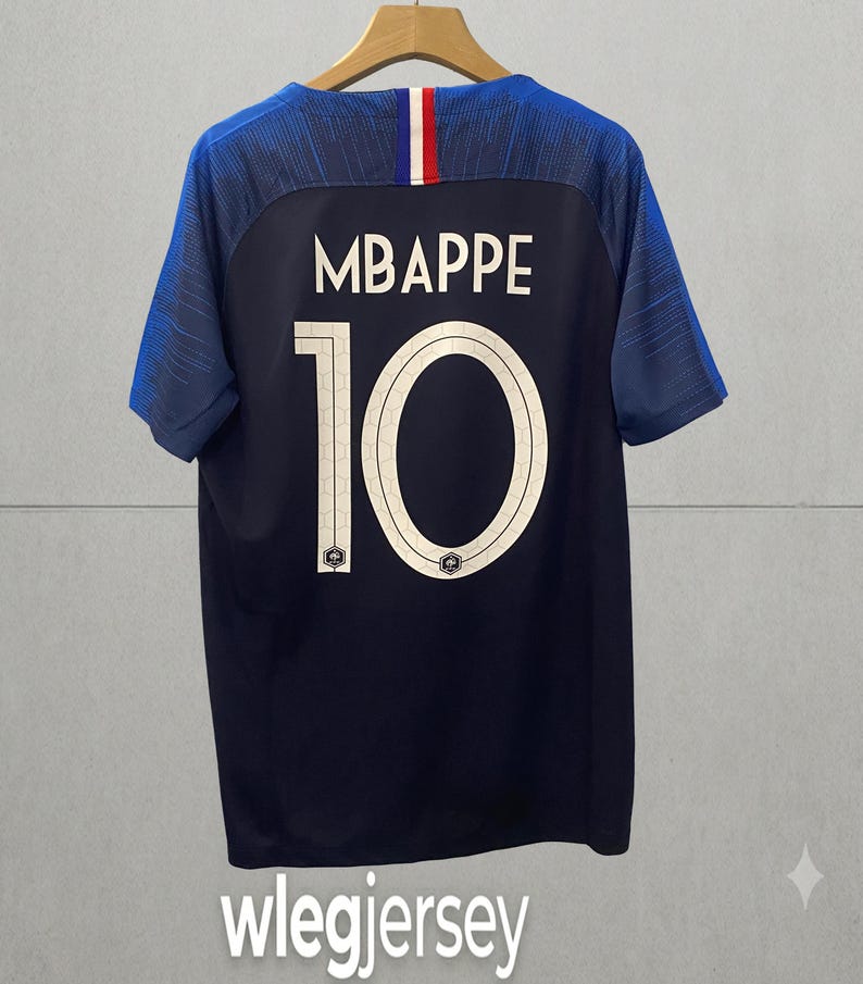 May include: A dark blue short-sleeved sports jersey featuring the name MBAPPE and the number 10 in white on the back. The collar area includes a vertical blue, white, and red stripe detail. The sleeves have a textured pattern in shades of blue.