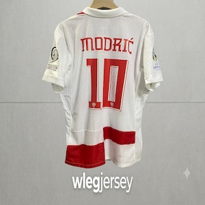 Pu&ograve; includere: Una maglia sportiva bianca a maniche corte con il nome MODRIĆ e il numero 10 in lettere rosse sul retro. La maglia presenta dettagli rossi sul bordo inferiore e stemmi ufficiali del torneo su entrambe le maniche, appesa a una gruccia.