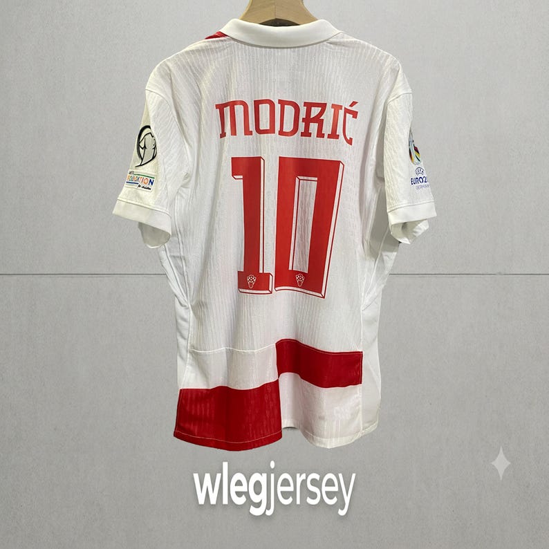 Pu&ograve; includere: Una maglia sportiva bianca a maniche corte con il nome MODRIĆ e il numero 10 in lettere rosse sul retro. La maglia presenta dettagli rossi sul bordo inferiore e stemmi ufficiali del torneo su entrambe le maniche, appesa a una gruccia.