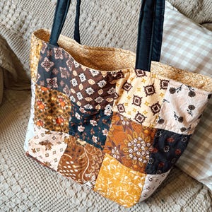 Könnte beinhalten: Eine gesteppte Tragetasche mit einem Patchwork-Design aus verschiedenen Stoffquadraten in Braun-, Creme- und Schwarztönen. Die Tasche hat zwei schwarze Stoffträger und ein gemustertes Innenfutter, das an der oberen Öffnung der Tasche sichtbar ist.