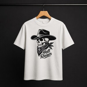 May include: A white short-sleeve t-shirt featuring a black graphic print of a skull wearing a cowboy hat and a patterned bandana. The text "Hell Raiser" is printed in a stylized black font directly below the bandana on the front of the shirt.