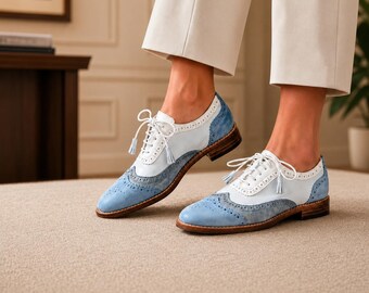 May include: A pair of light blue and white oxford shoes featuring a classic wingtip brogue design with decorative perforations, white laces with tassels, and a low stacked wooden heel. The shoes are worn with cropped cream-colored trousers.