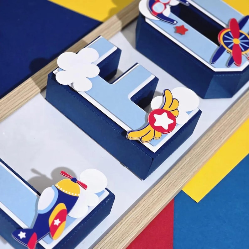 K&ouml;nnte beinhalten: Dekorative marineblaue und hellblaue Boxen in Form der Buchstaben "L", "E" und "O". Jede Box ist mit Flugzeug- und Wolkenausschnitten verziert. Die Boxen sind auf einer wei&szlig;en Oberfl&auml;che mit einem Holzrahmen angeordnet, mit gelben, roten und blauen Hintergr&uuml;nden.