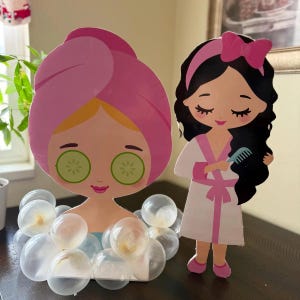 May include: Two cardboard cutouts of girls dressed in spa attire. One girl has a pink towel on her head and cucumber slices on her eyes. The other girl has a pink robe and is brushing her hair. Both girls have large eyes and are smiling. The girl with the towel is surrounded by white balloons.