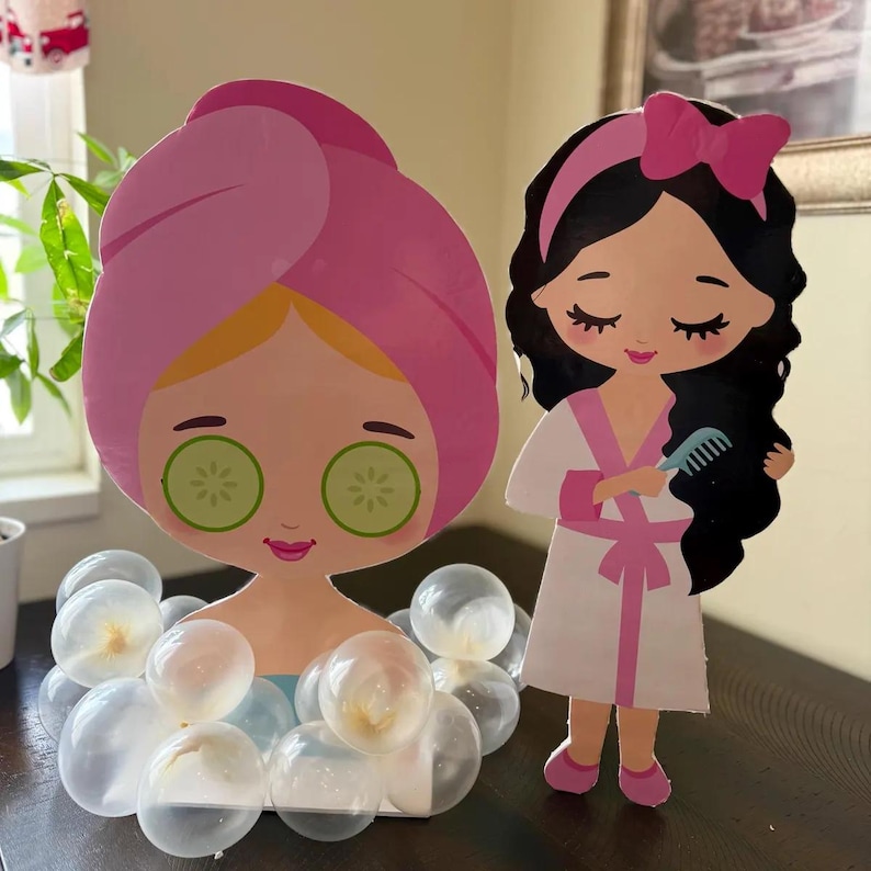 May include: Two cardboard cutouts of girls dressed in spa attire. One girl has a pink towel on her head and cucumber slices on her eyes. The other girl has a pink robe and is brushing her hair. Both girls have large eyes and are smiling. The girl with the towel is surrounded by white balloons.