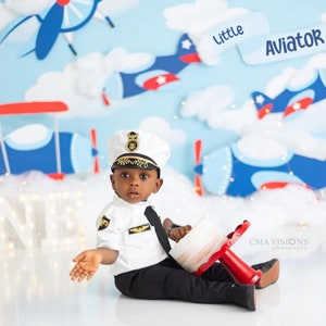 K&ouml;nnte beinhalten: Ein Kind in einer wei&szlig;en Pilotenuniform mit M&uuml;tze und Krawatte sitzt auf dem Boden. Der Hintergrund zeigt eine Szene mit Flugzeugen und Wolken. Das Kind h&auml;lt einen rot-wei&szlig;en Gegenstand. Der Text "Little Aviator" ist sichtbar.