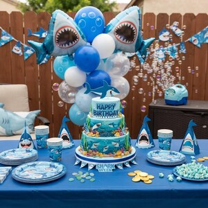 May include: A three-tiered birthday cake decorated with shark imagery and the text Happy Birthday, surrounded by shark-themed party supplies including plates, cups, party hats, and a balloon arch featuring blue and white balloons and shark foil balloons.