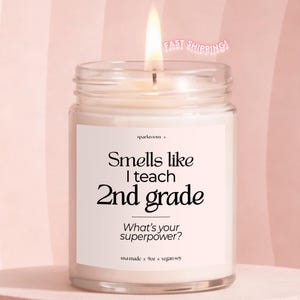 May include: A lit candle in a clear glass jar with a white label featuring the black text: Smells like I teach 2nd grade. Below this, the label reads: What's your superpower? The bottom of the label includes the text: usa made 9oz vegan soy.