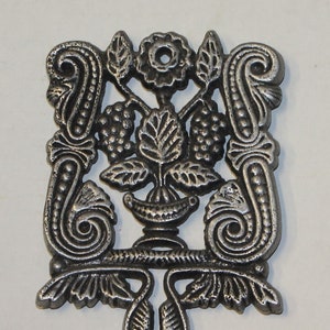 Puede incluir: Un salvamanteles de hierro fundido plateado con un diseño floral. El salvamanteles presenta una flor central con hojas y bayas. El salvamanteles mide aproximadamente 7,6 cm de alto.