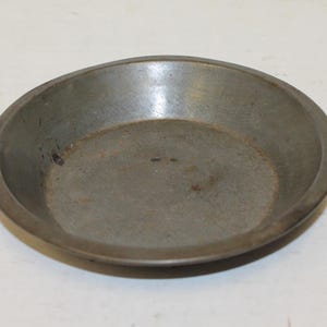May include: A small, round, metal pie dish with a slightly raised rim. The dish has a dull, grey colour with some darker spots and a slightly rough texture. The interior is smooth and shows signs of use.