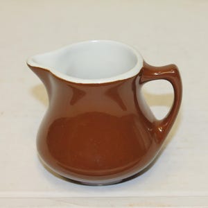 May include: A small, glossy brown ceramic creamer pitcher with a smooth, rounded body, a curved handle on the right side, and a white interior. The pitcher features a distinct pouring spout on the left side, designed for serving liquids like cream.