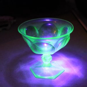 Op de afbeelding: Een heldergroen glazen dessertgerecht met een zeshoekige voet en een decoratieve steel. De kom heeft een geschulpte rand en een geometrisch ontwerp. Het glas lijkt te gloeien onder zwart licht.