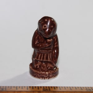 May include: A small, brown ceramic figurine of a monkey in a seated position. The monkey is wearing a robe-like garment and has its hands clasped in front. The figurine is on a decorative base.