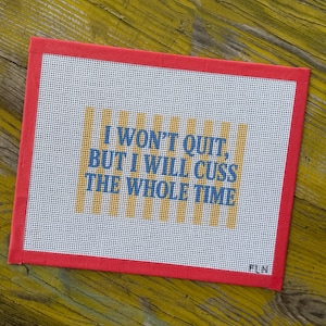 May include: A rectangular needlepoint canvas featuring a red border and a white grid background. In the center, yellow vertical stripes sit behind the blue text that reads: I WON'T QUIT, BUT I WILL CUSS THE WHOLE TIME. The canvas has a small signature.