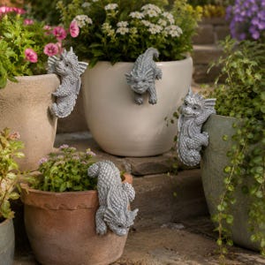 Peut inclure: Quatre figurines de dragon en pierre grise sont perchées sur les bords de divers pots de fleurs en céramique. Chaque dragon présente des écailles texturées, de petites ailes et un visage souriant, positionné pour grimper sur le rebord.