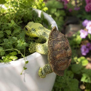 May include: A decorative turtle figurine with a textured brown shell and a light green, speckled body. The turtle is positioned to hang over the edge of a smooth white planter filled with green leafy plants, showing its head and limbs.