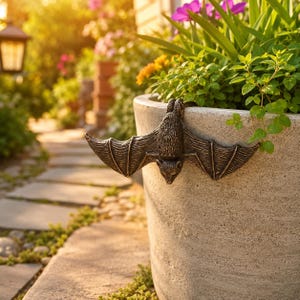 Peut inclure: Une figurine de chauve-souris de couleur bronze foncé conçue pour être suspendue au bord d'un grand pot de fleurs en pierre claire. La chauve-souris présente des ailes texturées et un visage détaillé, positionnée horizontalement sur le rebord.